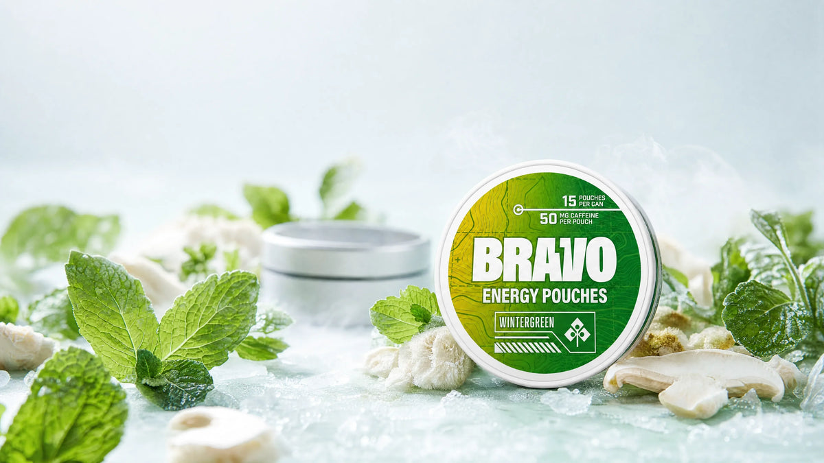 Boost Pouches review hero: Bravo Wintergreen caffeine pouch tin in sharp focus with mint leaves and mushroom accents, unbranded soft-focus competitor tin in background